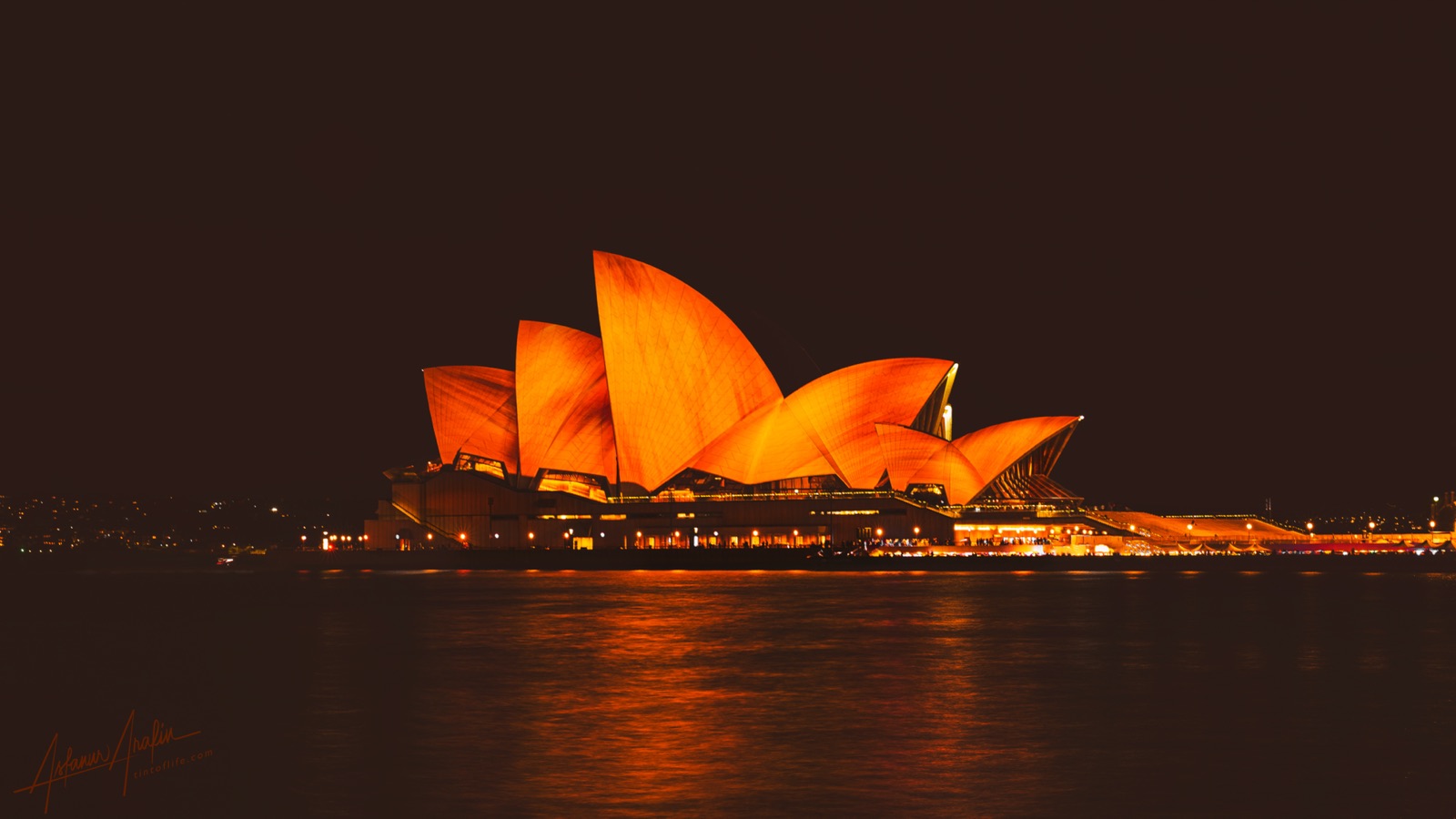 Sydney Opera House lit deep orange at night, reflected across the harbour